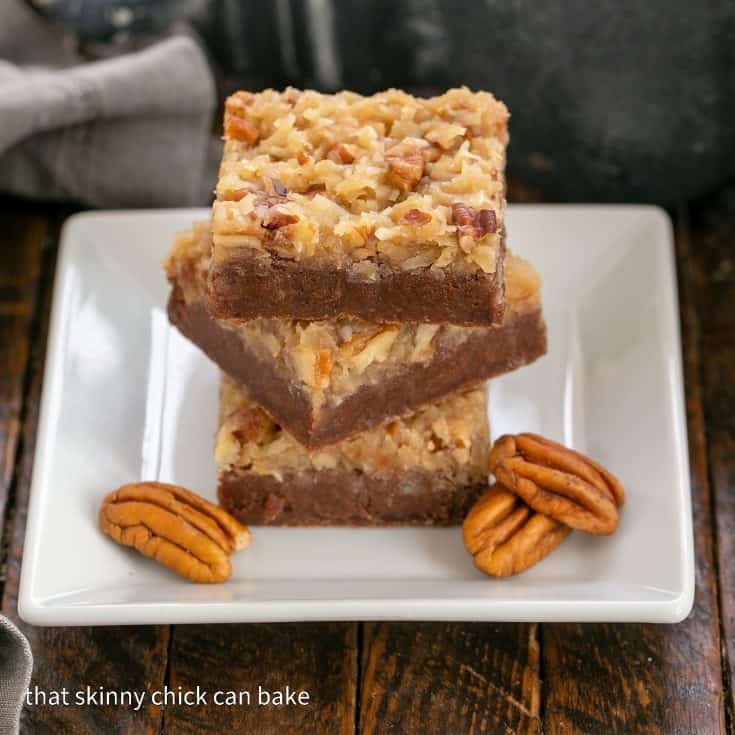 3 German chocolate brownies stacked on a square white plate with 3 pecans to garnish