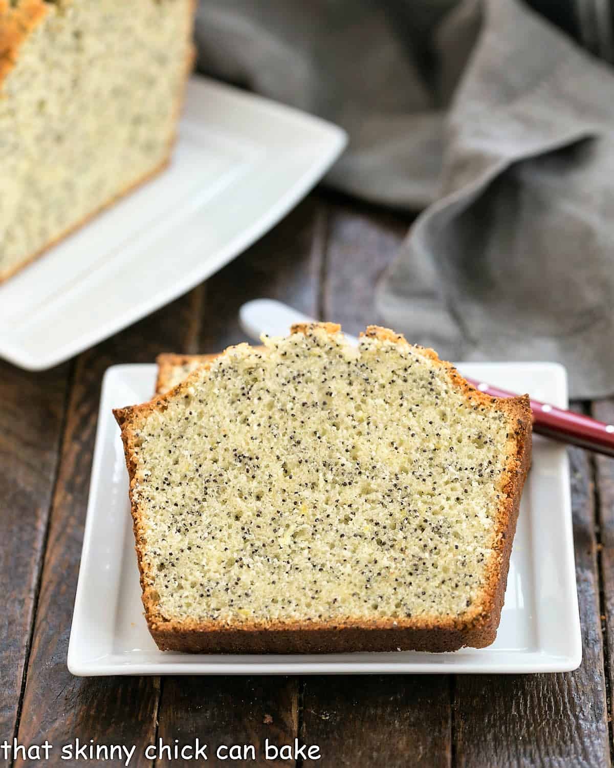 Two slices of poppy seed tea cake on a square white plate with a red handle knife in front of the rest of the loaf on a ceramic tray.