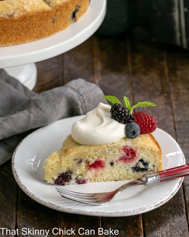 Slice of berry cake recipe with lemon whipped cream on a white plate in front of a cake stand