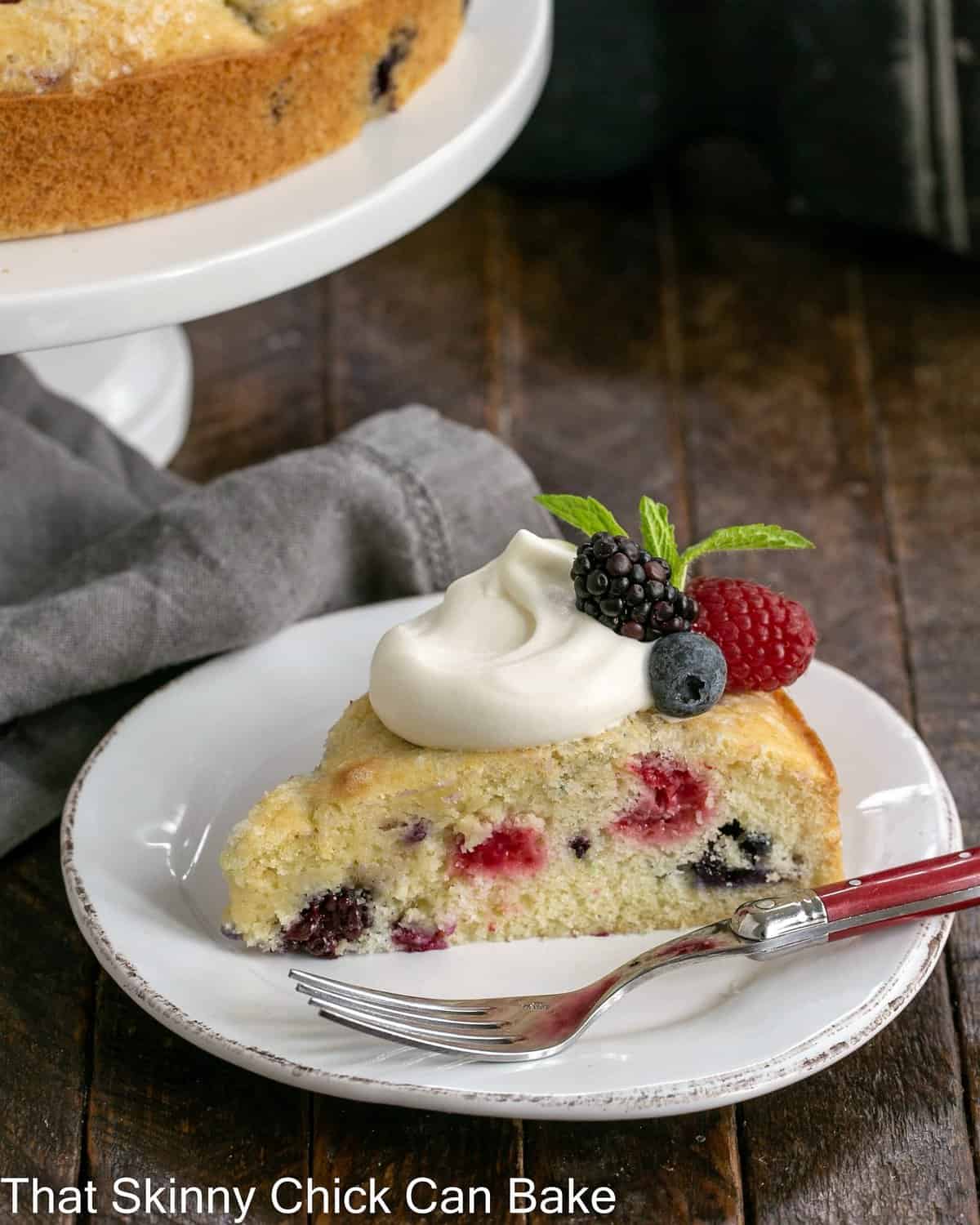 Slice of berry cake recipe with lemon whipped cream on a white plate in front of a cake stand.