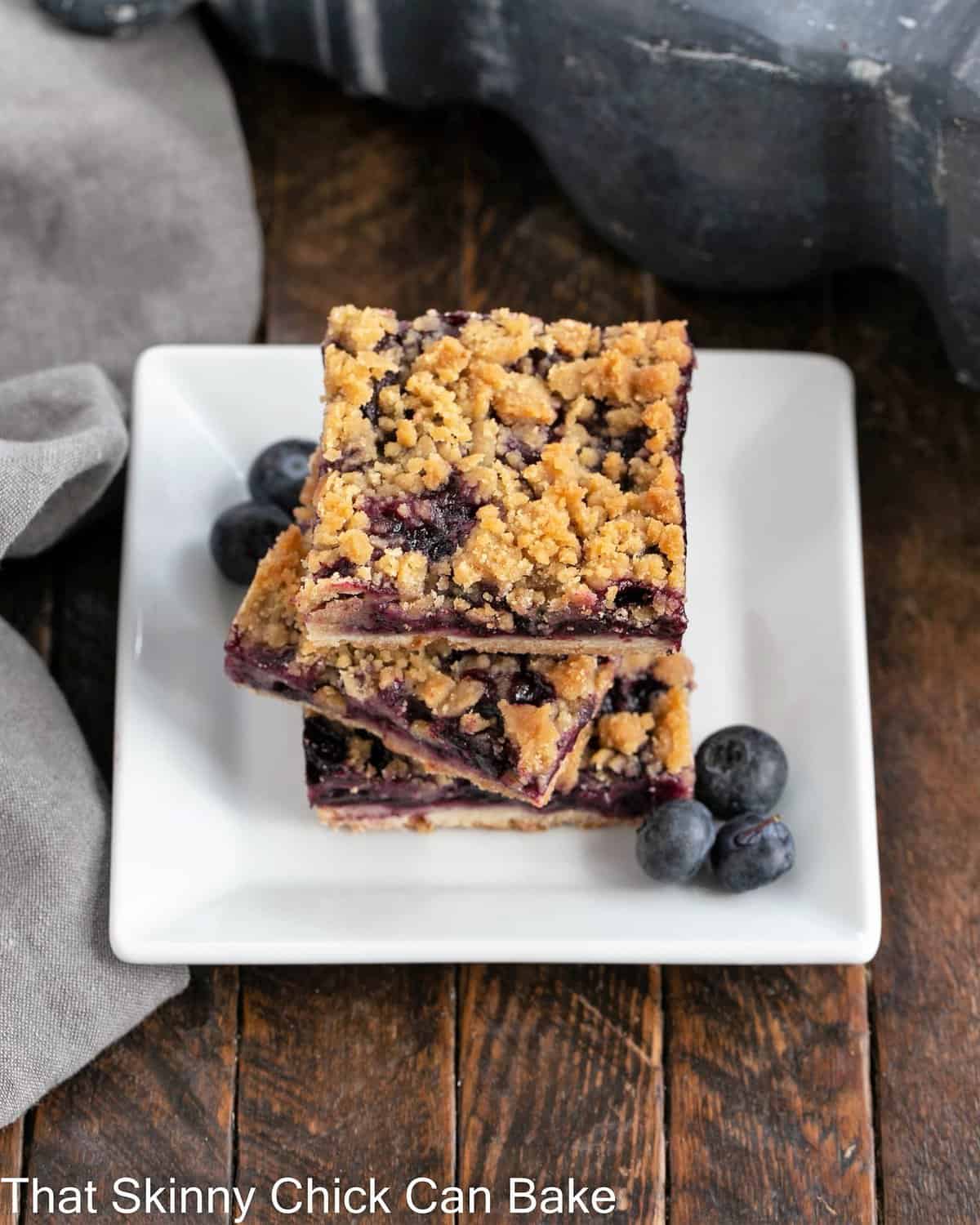 3 blueberry bars stacked on a square white dessert plate with fresh blueberries to garnish.