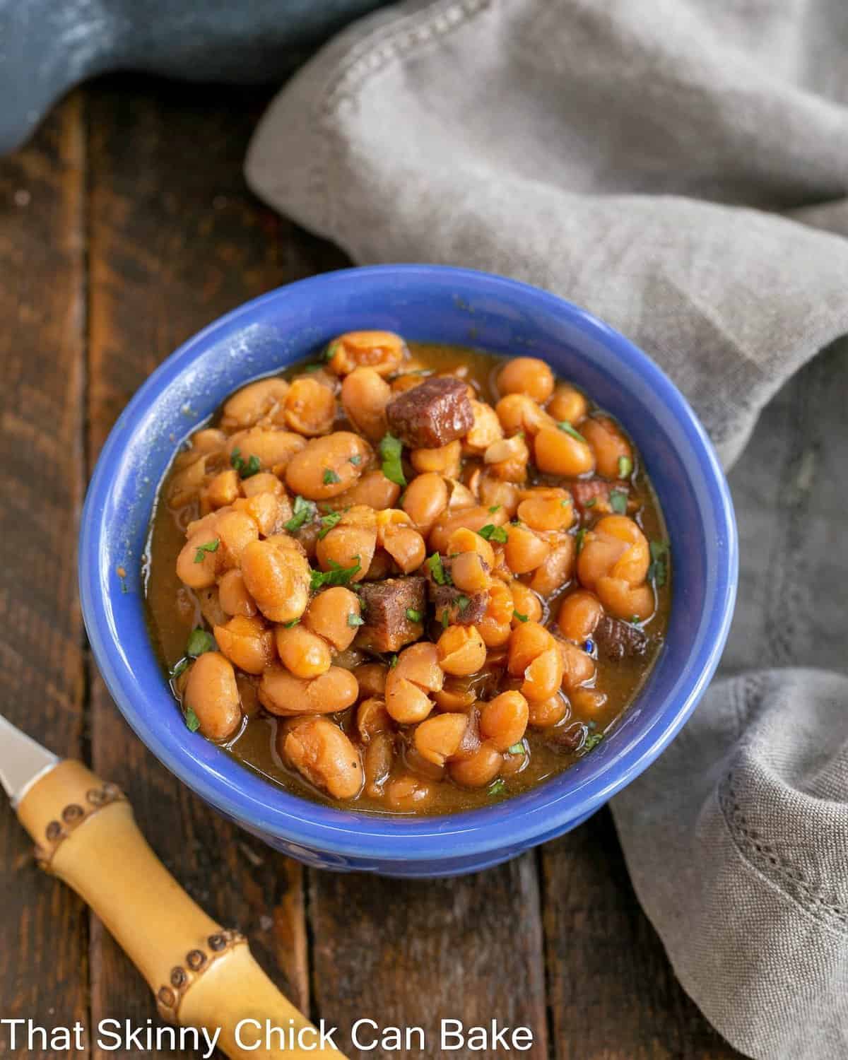 homemade baked beans in a blue bowl with a bamboo fork to the lower left.