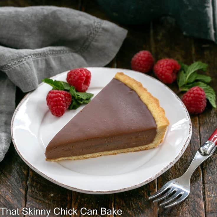 A wedge of milk chocolate tart on a white dessert plate with raspberry garnish and a red handle fork