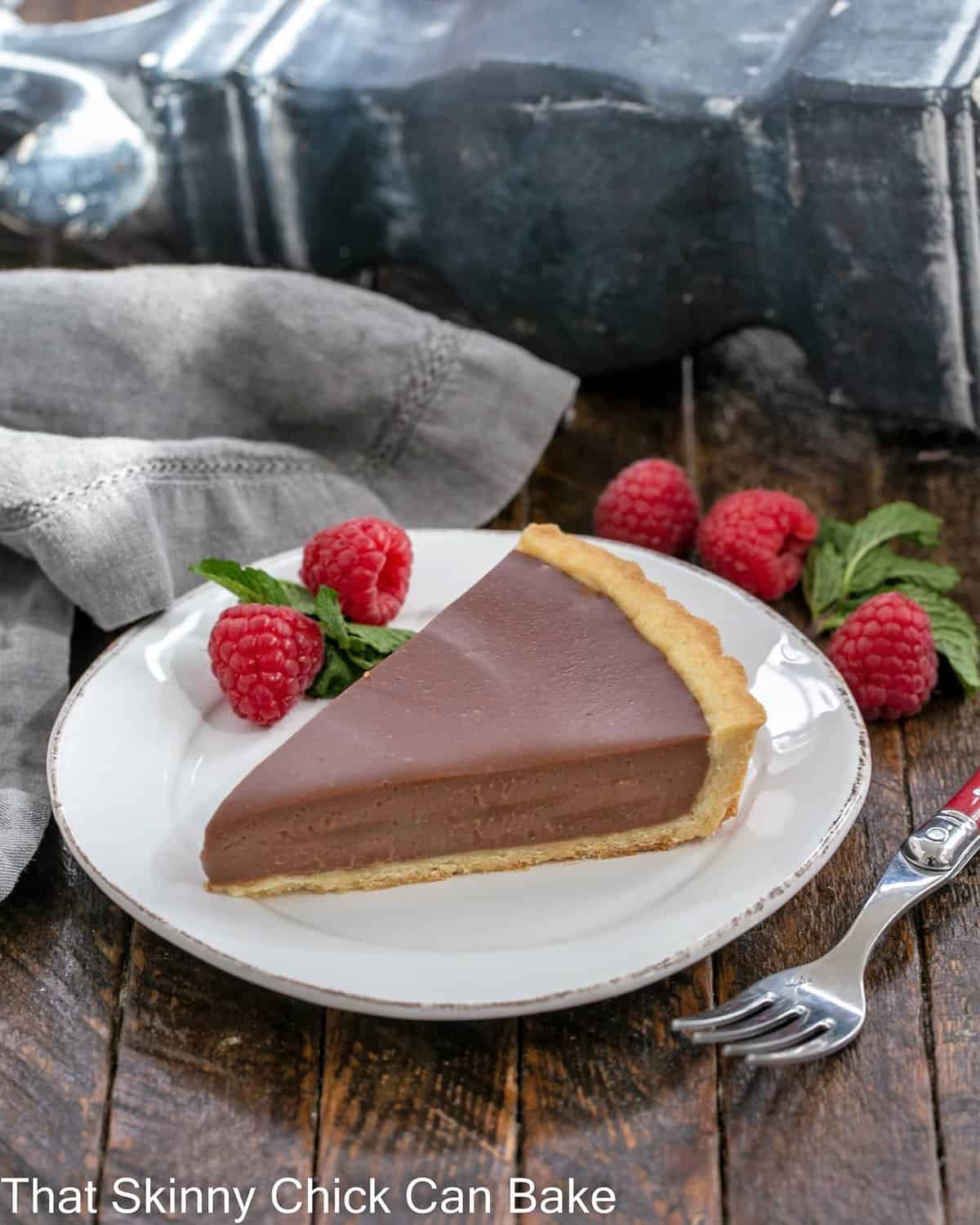 A slice of milk chocolate tart on a white plate with a raspberry and mint garnish.