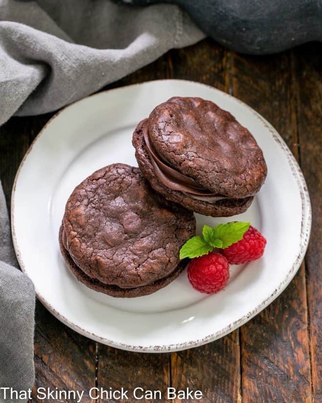 Overhead view of two chocolate sandwich cookies on a white dessert plate with 2 raspberries and a sprig of mint
