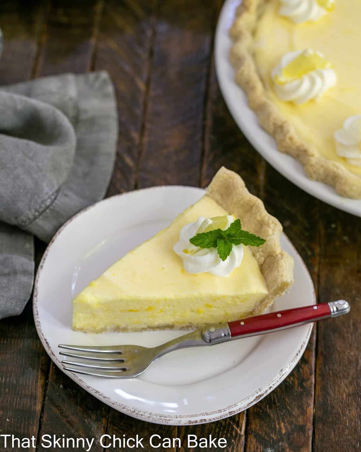 Slice of lemon chiffon pie on a white plate with a red handle fork and whole pie in the background.