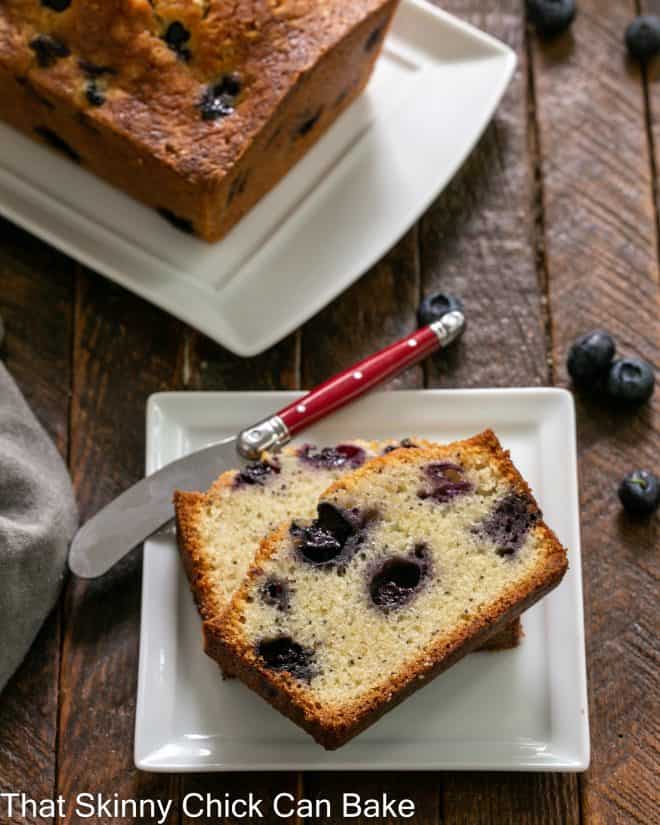 Two slices of blueberry bread on a white plate in front of a loaf on a serving tray.