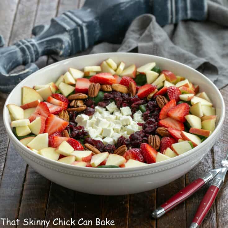 Spinach salad with strawberries and feta in a white ceramic salad bowl and 2 serving utensils.