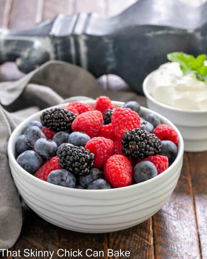 A white bowl of mixed berries in front of a bowl of chantilly cream.