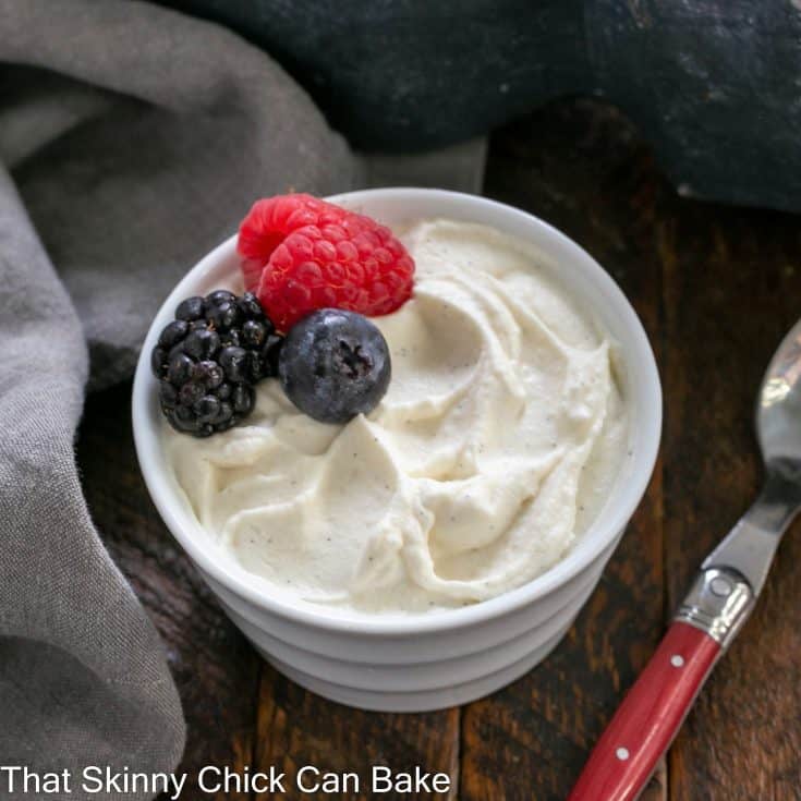 Overhead view of a white ramekin filled with Chantilly Cream garnished with 3 fresh berries next to a red handled spoon