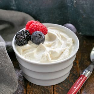 Crème Chantilly in a white ramekin topped with 3 berries next to a red handled spoon