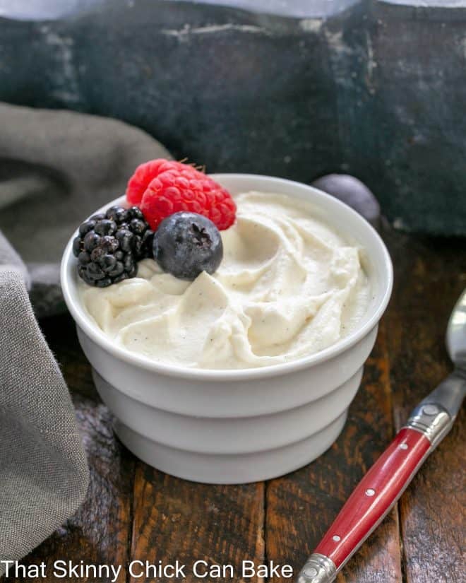 Crème Chantilly in a white ramekin topped with 3 berries next to a red handled spoon.