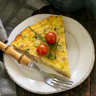 Overhead view of a slice of zucchini quiche on a small white plate with a bamboo handled fork