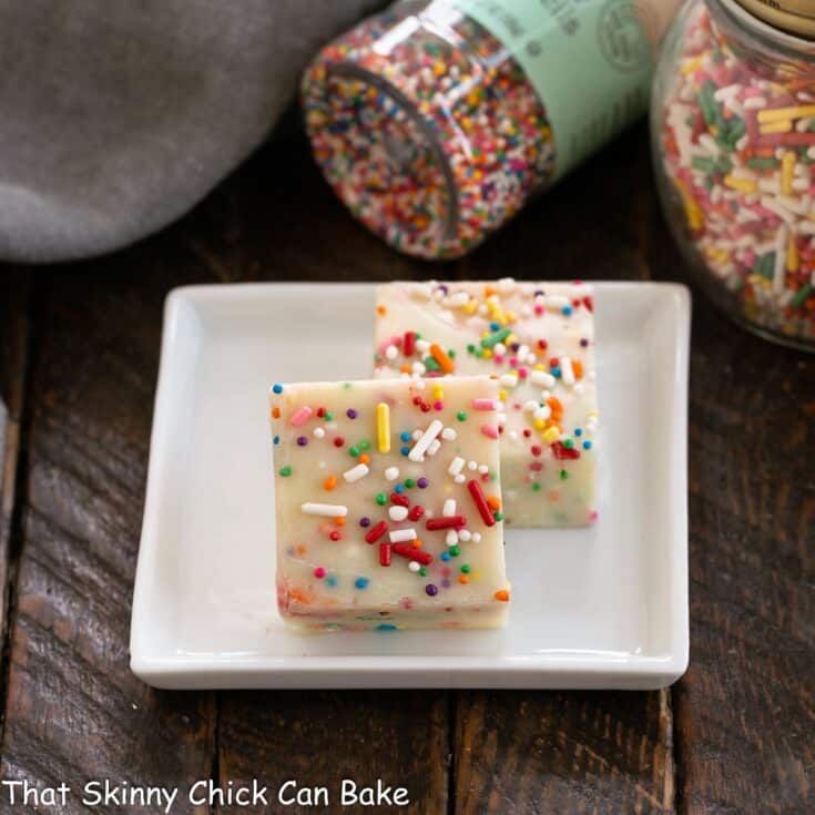 2 squares of white chocolate fudge on a white plate with jars of sprinkles.