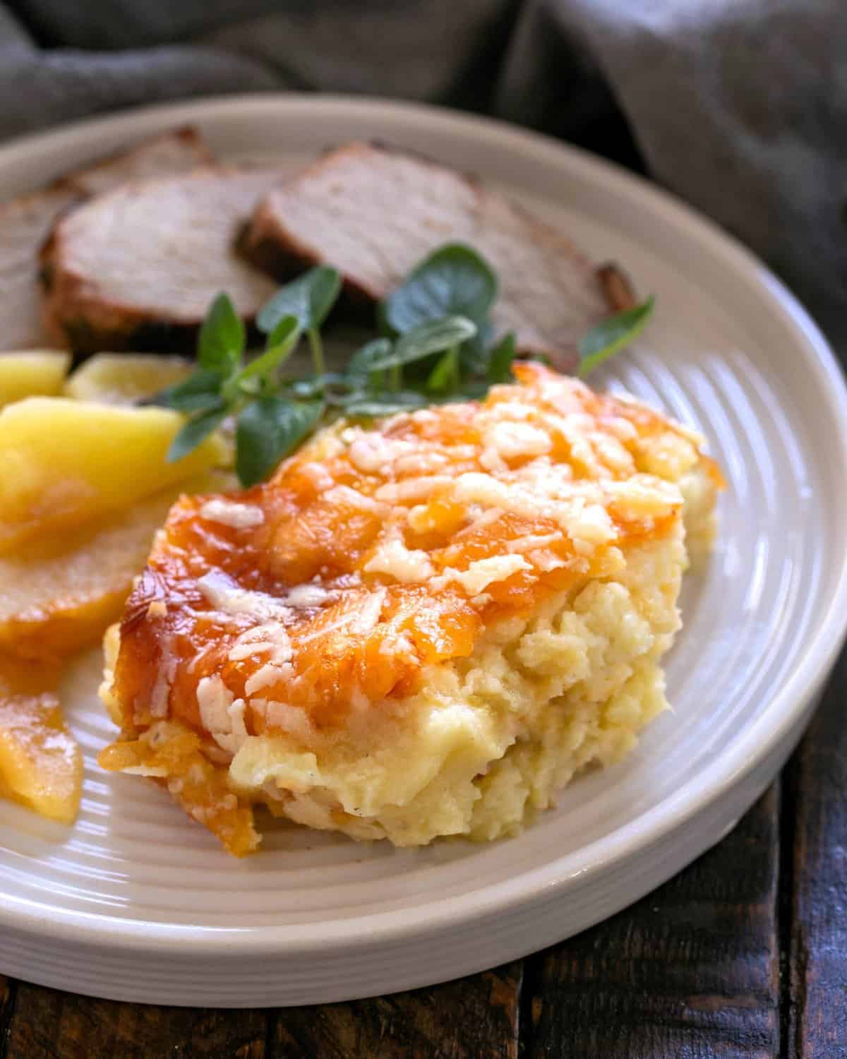 Serving of potato casserole on a white dinner plate with pork and apples.