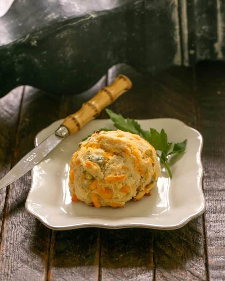 A cheddar biscuit on a white plate with a sprig of parsley and a knife.