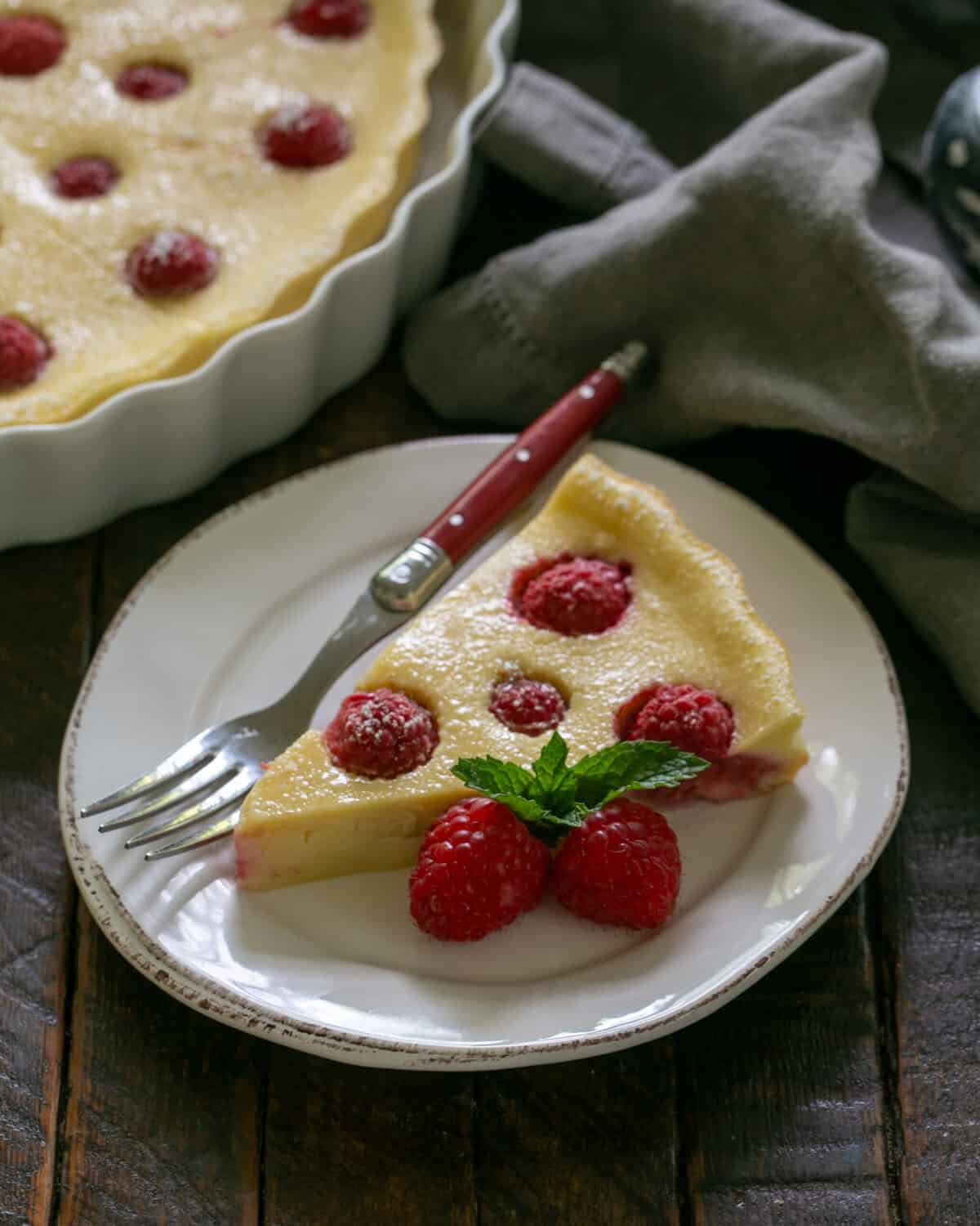 Slice of a raspberry clafoutis with a red handled fork on a white dessert plate with the serving dish in the background.